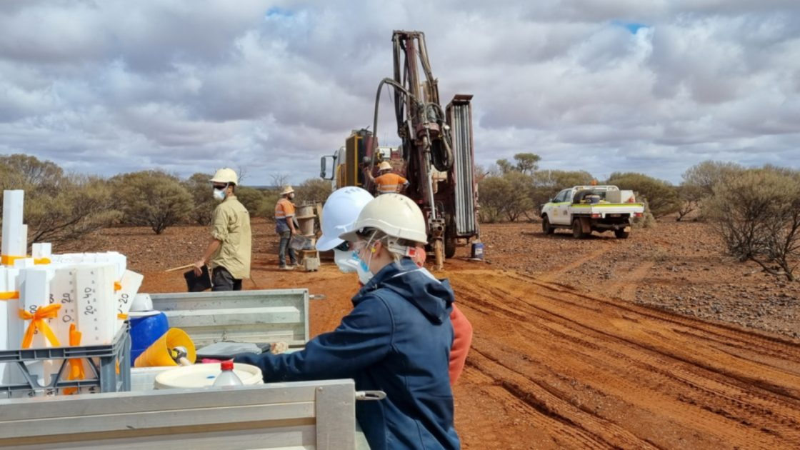 Geological field workers wearing hard hats and protective masks organizing core sample trays on a truck bed near an active drilling rig at the Great Boulder Resources Peak Hill Gold Project in Western Australia
