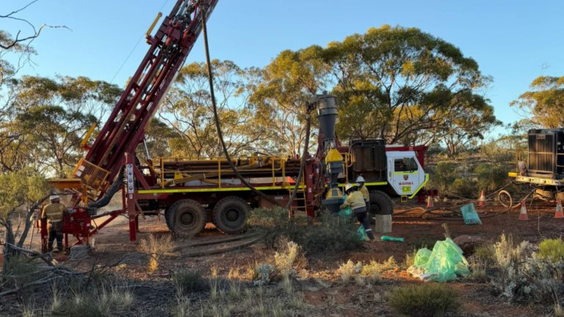 A truck-mounted exploration drill rig with a raised red mast operating in an Australian bush setting, with workers in high-visibility gear collecting samples in green bags on red dirt ground at Evergold Minerals' Rabbit Warren gold discovery in Western Australia